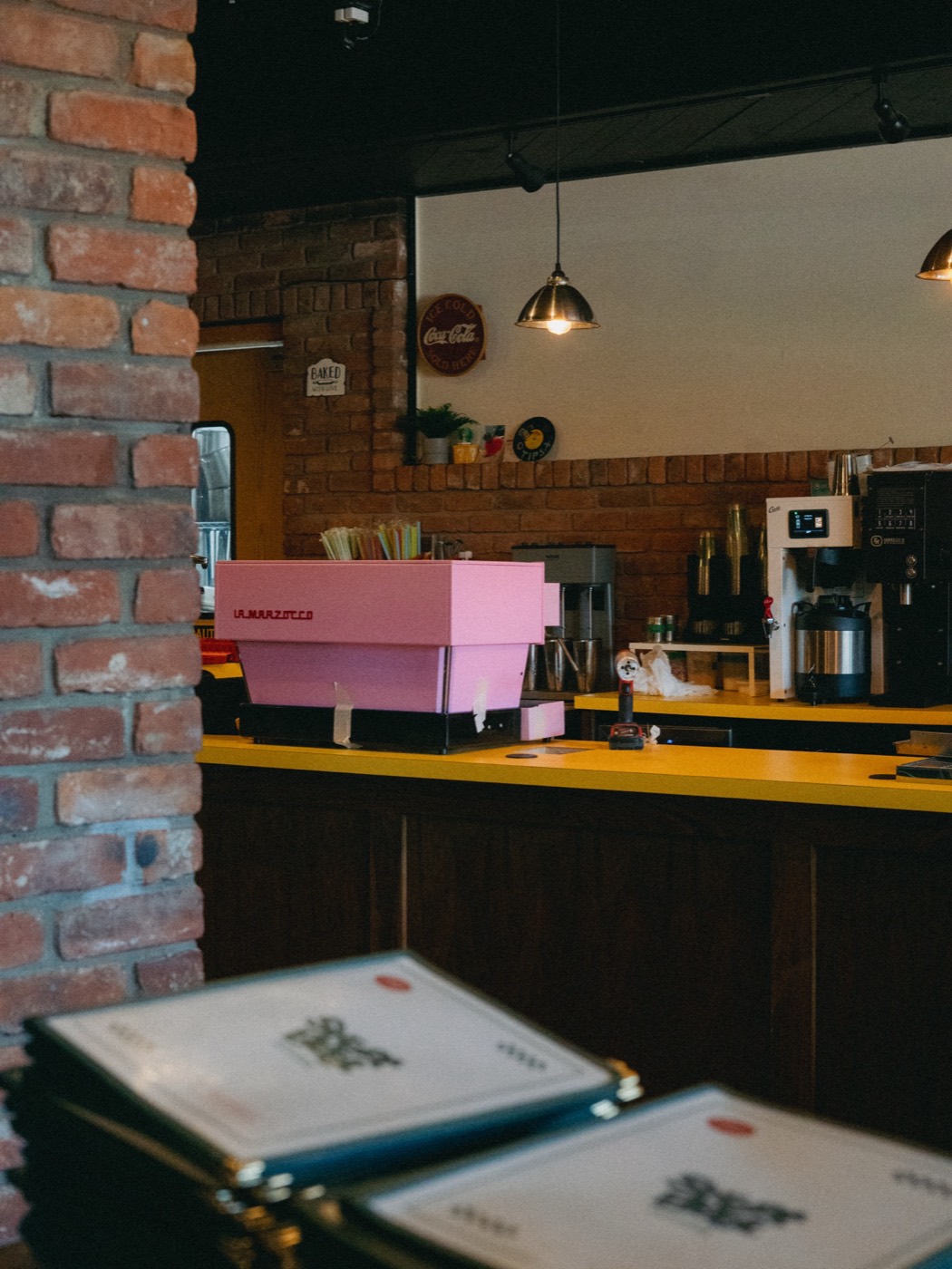A pink La Marzocco espresso machine on a yellow counter inside a diner, viewed through a brick passage