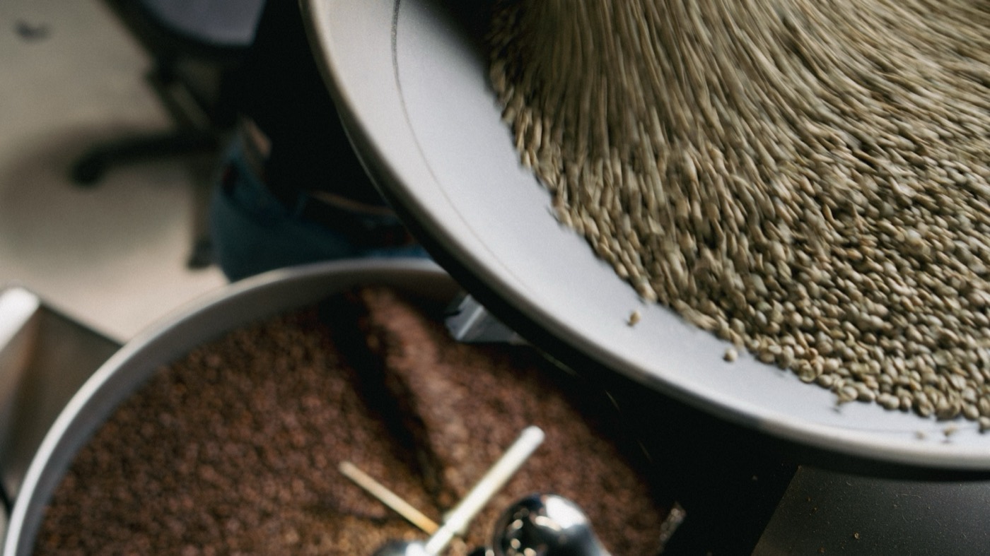 Green coffee beans being poured into the roaster alongside a tray of just-roasted beans at Coffeehaus